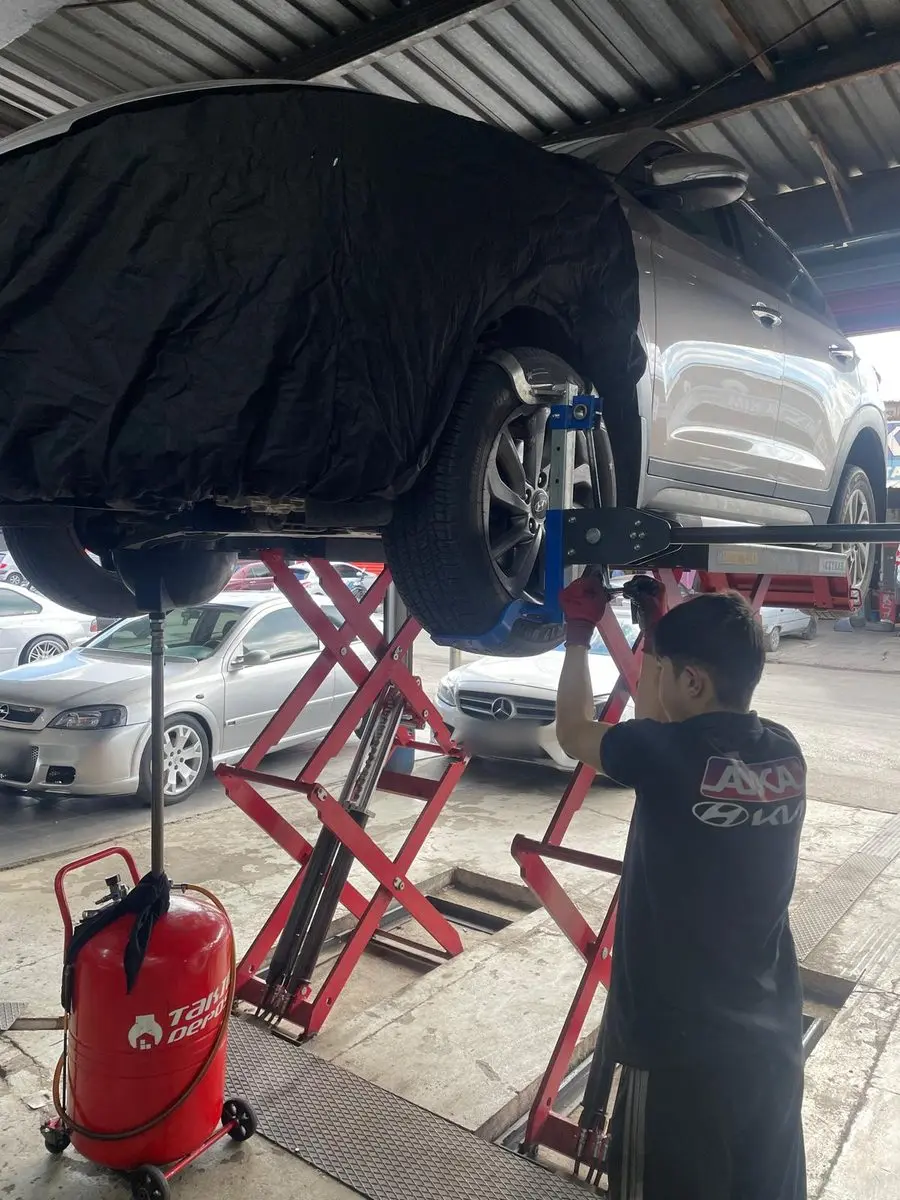 Örtüyle korunan, lift üzerinde kaldırılmış aracın tekerleğinde çalışan usta; yanda yağ boşaltma kabı